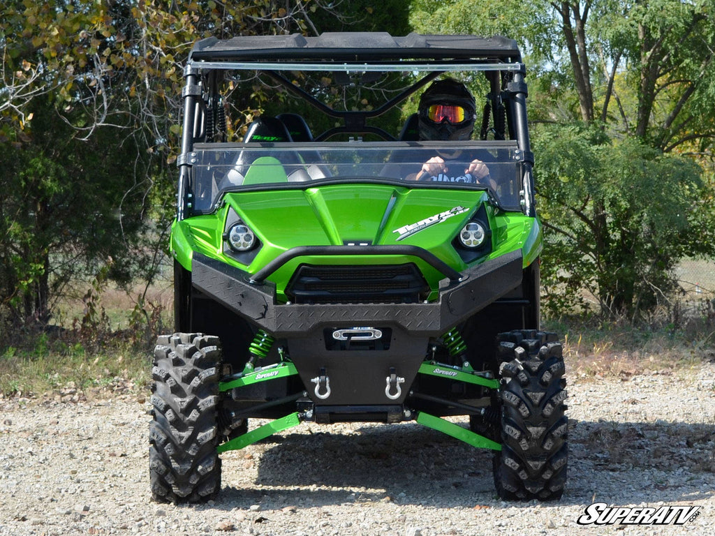 A Kawasaki Teryx fitted with a Flip Windshield is parked on a gravel path, surrounded by trees. Two helmeted riders sit inside the bright green off-road vehicle, which has large rugged tires. The SuperATV logo appears in the bottom right corner.