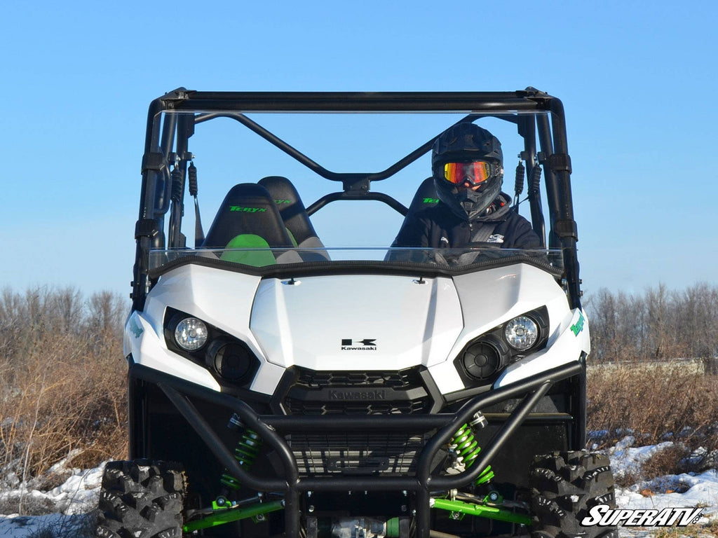 A person wearing a helmet and goggles drives a white Kawasaki Teryx 800 equipped with a Full Windshield through a grassy field under a clear blue sky. The ATV features green springs, rugged tires, and SuperATV branding in the bottom right corner.