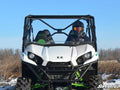 A person wearing a helmet and goggles drives a white Kawasaki Teryx 800 equipped with a Full Windshield through a grassy field under a clear blue sky. The ATV features green springs, rugged tires, and SuperATV branding in the bottom right corner.