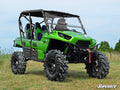 A Kawasaki Teryx equipped with a 4 Portal Gear Lift sits on grass. This bright green, four-seat side-by-side features large rugged tires, a black roll cage, windshield, roof, bucket seats with seat belts, and SuperATV branding in the lower right.