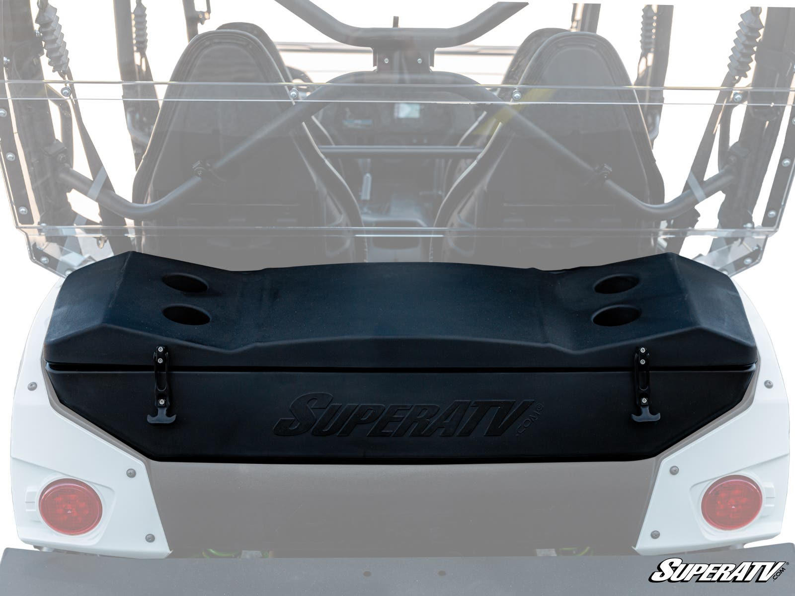 A Kawasaki Teryx 4 Cargo Box in black is mounted to the rear of a white off-road UTV. The box features four cup holders on top, side latches, and displays the SuperATV logo. The image shows the UTV’s roll cage and seats.