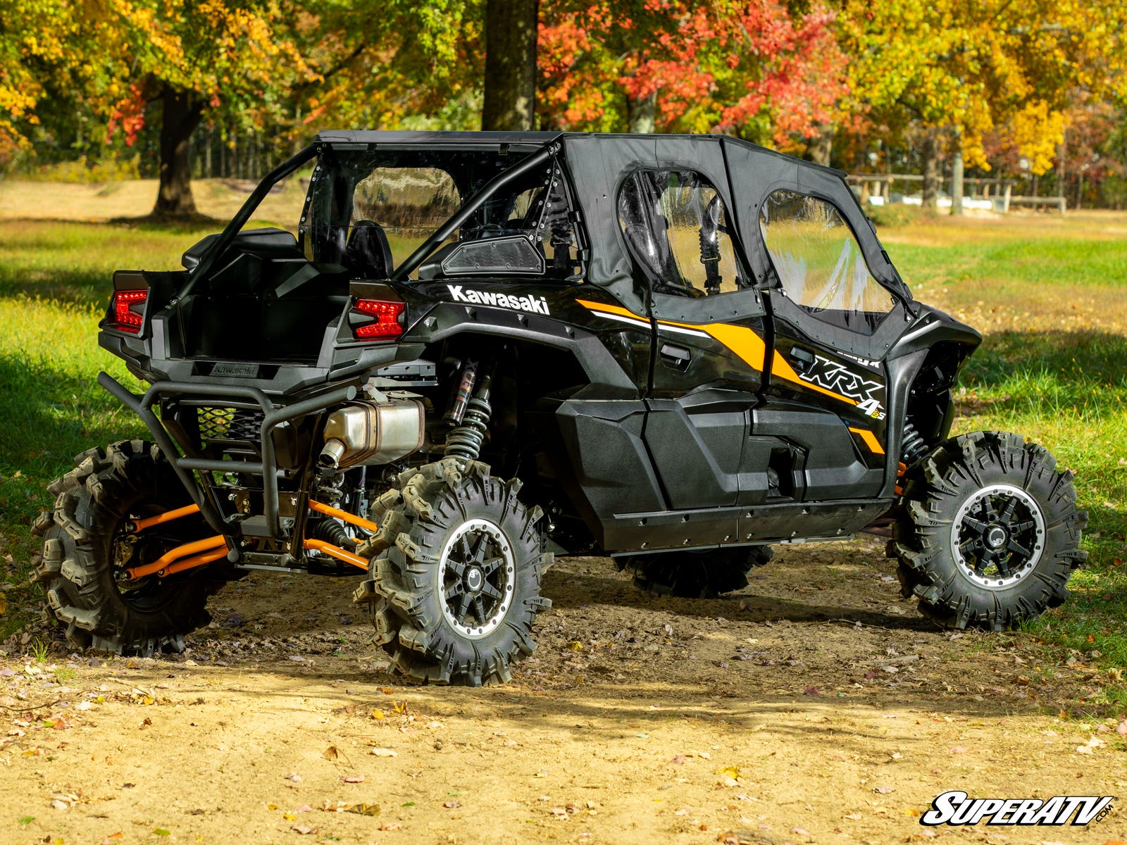 A black and yellow Kawasaki Teryx KRX 4 1000 with large rugged tires and Primal Soft Cab Enclosure Upper Doors is parked on a dirt path among green grass and autumn trees. SuperATV is watermarked on the image.