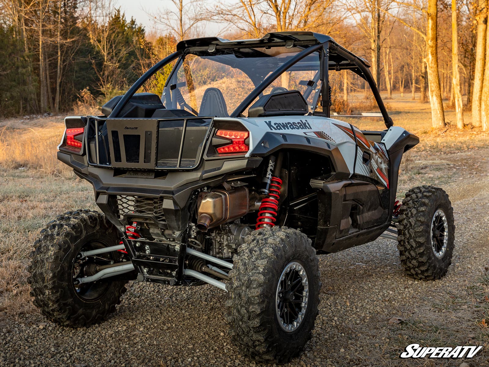 A Kawasaki Teryx KRX 1000 featuring a rear windshield is parked on a gravel path in a wooded area. The black and gray side-by-side has rugged tires, red coilover suspension, bucket seats, and displays the SuperATV logo in the bottom right corner.