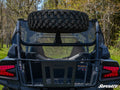 A rear view of a black off-road vehicle features the Kawasaki Teryx KRX 1000 Spare Tire Carrier mounted above the back seats. Red taillights show, and it’s parked outdoors with trees and grass visible. The “SuperATV” logo appears in the bottom right corne