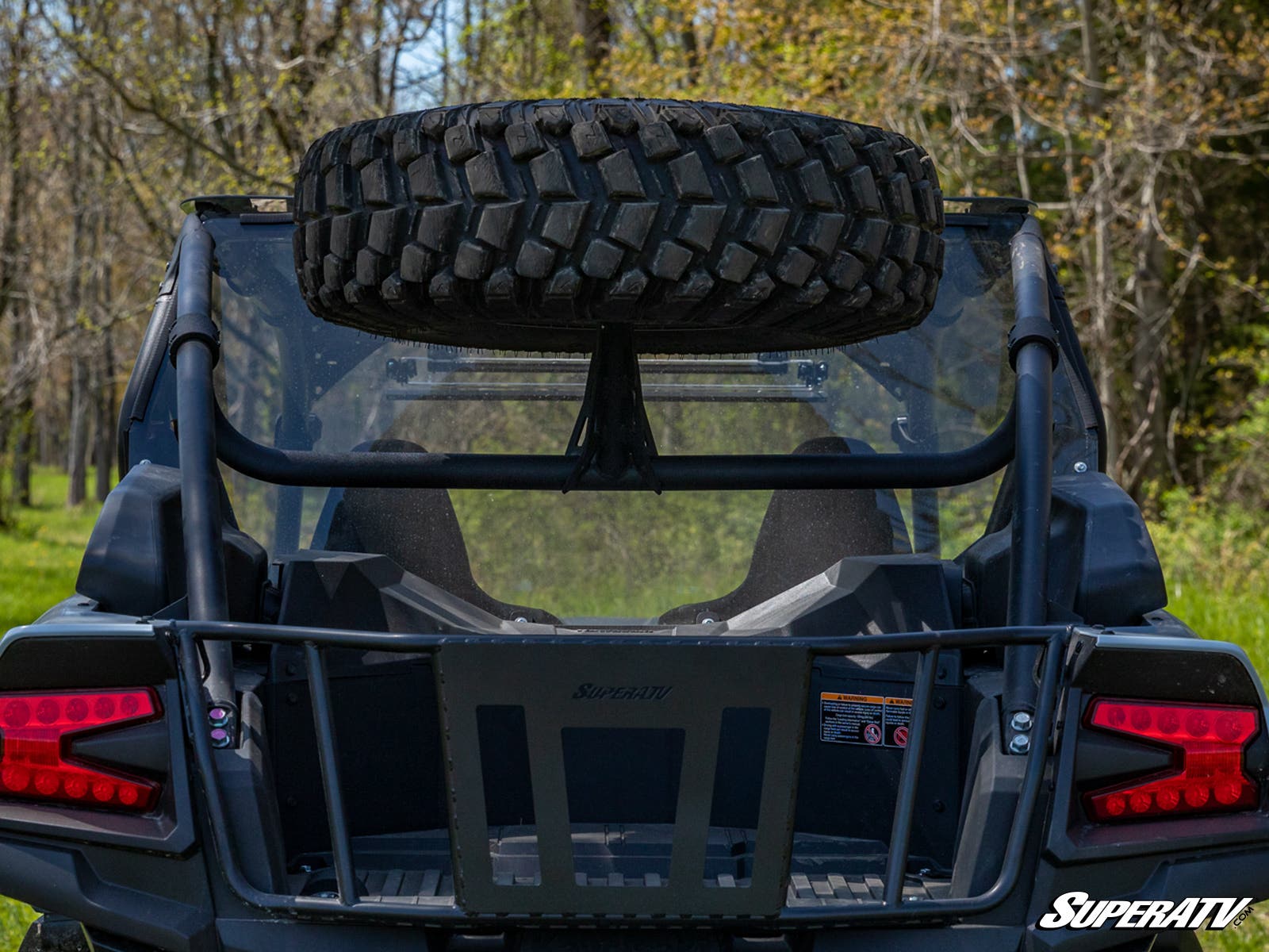 A rear view of a black off-road vehicle features the Kawasaki Teryx KRX 1000 Spare Tire Carrier mounted above the back seats. Red taillights show, and it’s parked outdoors with trees and grass visible. The “SuperATV” logo appears in the bottom right corne