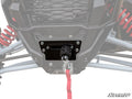 Close-up of the Kawasaki Teryx KRX Winch Mounting Plate installed on an off-road vehicle. A metal-hook winch with a red strap, red suspension springs, and gray frame parts are visible. The SuperATV logo appears in the bottom right corner.