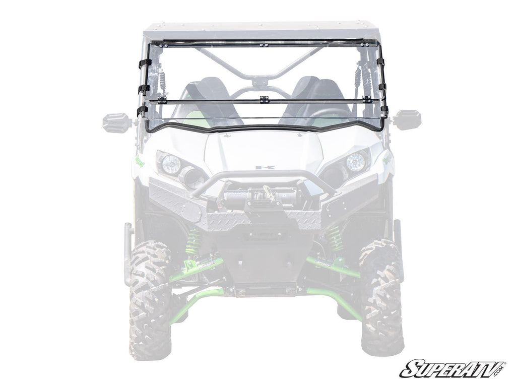 A full-front view of a Kawasaki Teryx S with a Flip Down Windshield installed. The clear, segmented polycarbonate windshield features a center hinge, wipers, metal brackets, and the “SuperATV” logo in the bottom right corner.