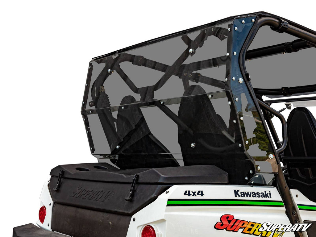 A close-up of a Kawasaki Teryx 4 S Rear Windshield on a white 4x4 side-by-side shows its tinted, securely attached rear windshield, black roll cage, and two seats; the SuperATV logo appears in the bottom right corner.