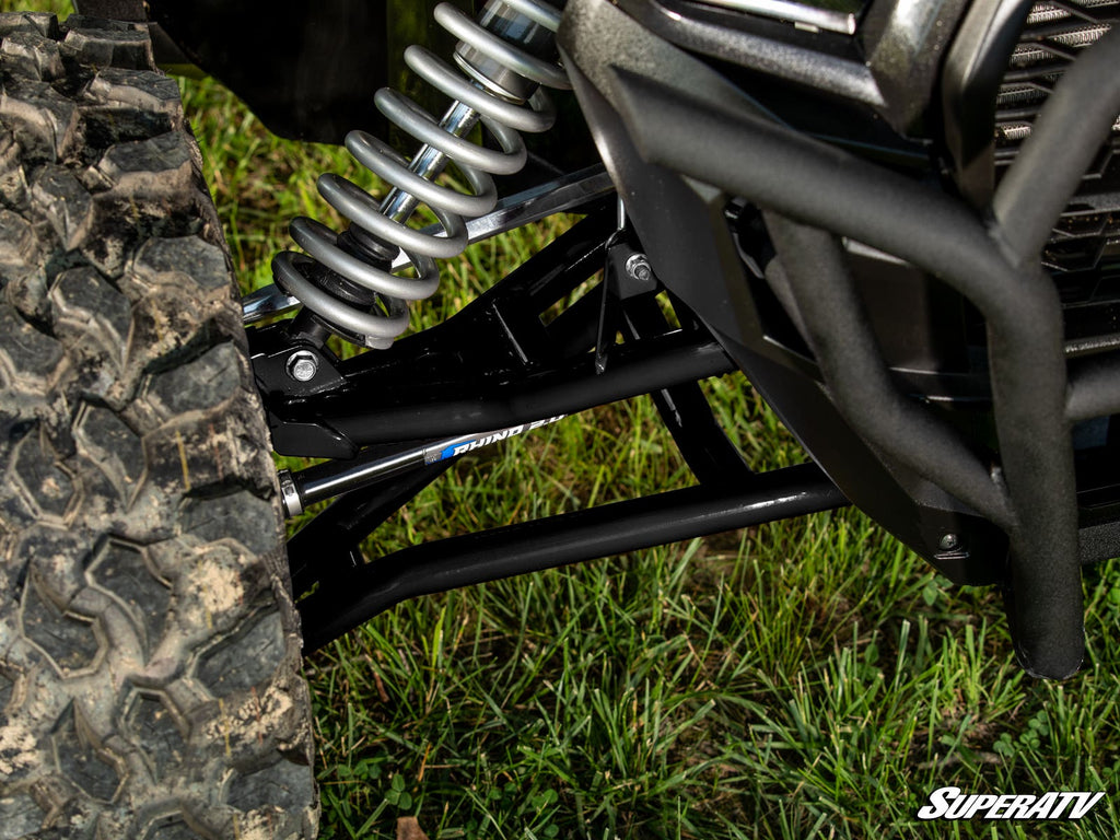 Close-up of a Kawasaki Teryx KRX 1000 Atlas Pro 1.5 Forward Offset A-Arms with a large coilover shock, black control arms, and a rugged mud tire. The SuperATV logo appears in the bottom right. Vehicle is parked on grass.