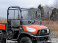 A Kubota RTV with a Scratch-Resistant Full Windshield and mesh back window is parked on a dirt path. Featuring rugged tires and two black seats, its surrounded by leafless trees and dry grass under a partly cloudy sky.