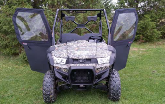 The Kymco UXV 450 with Convertible Doors sits on green grass with both side doors open, showing clear panel windows and rugged tires. Its camouflage pattern blends into the evergreen trees in the background.