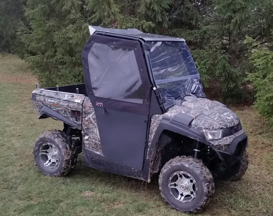 A Kymco UXV 450 Cab Kit with Aluminum Roof, featuring a camouflage pattern, clear windshield, partial soft doors, large off-road tires, and rear cargo bed, is parked on grass near tall evergreen trees.