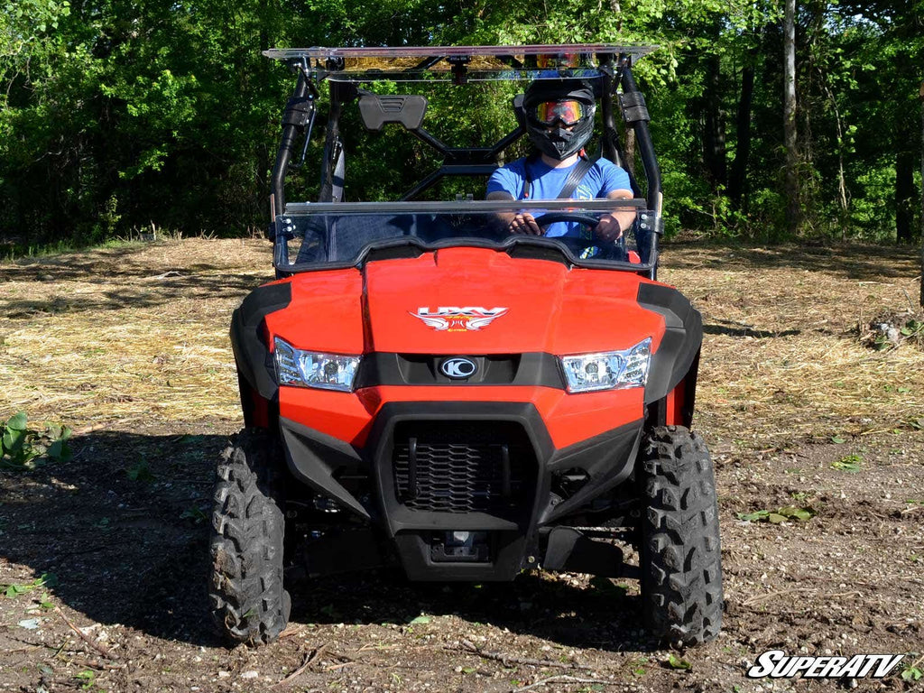 A person in a helmet and blue shirt sits in a bright red off-road vehicle on rough terrain. The Kymco UXV 450 Flip Windshield is installed, with large tires, sturdy bumper, and trees in the background. SuperATV logo at lower right.