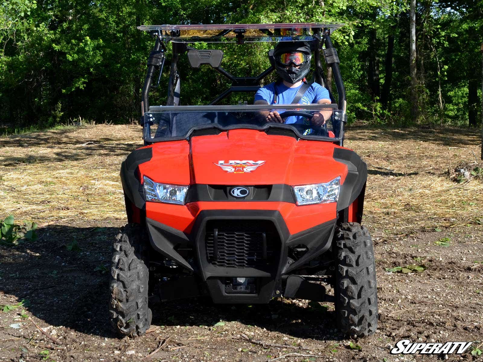 A person in a helmet and blue shirt sits in a bright red off-road vehicle on rough terrain. The Kymco UXV 450 Flip Windshield is installed, with large tires, sturdy bumper, and trees in the background. SuperATV logo at lower right.