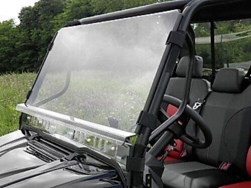A close-up of an Arctic Cat Prowler 700 Lexan Windshield in a black metal frame, with black and red front seats visible inside the off-road vehicle. Green foliage and grass can be seen in the background.