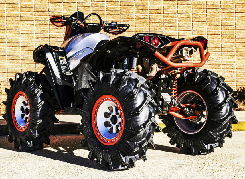 A black and silver ATV fitted with S3 Power Sports 2013-2018 Can-Am Renegade/Outlander +2 Stretched Hd Trailing Arms, oversized deep-tread tires, orange rims, and exposed suspension is parked before a tan brick wall.
