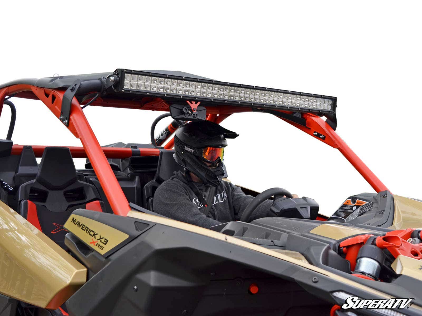 A person in a black helmet and goggles sits in the driver’s seat of a tan and red Can-Am Maverick X3 equipped with a large LED light bar mounted using the Can-Am Maverick X3 Light Bar Mounting Kit. The background is plain white.