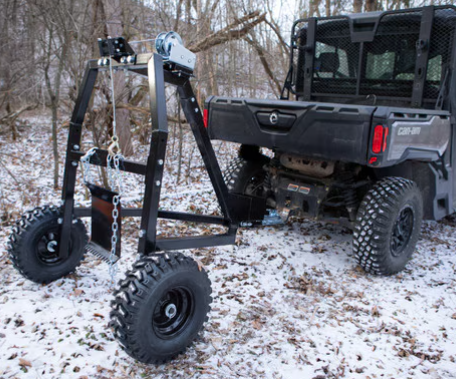 A black utility vehicle is parked in a snowy wooded area with a Log Hauler—a large, wheeled metal frame with a winch and chains—attached to its hitch, likely used for lifting or hauling heavy objects like logs or equipment.