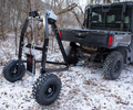 A black utility vehicle is parked in a snowy wooded area with a Log Hauler—a large, wheeled metal frame with a winch and chains—attached to its hitch, likely used for lifting or hauling heavy objects like logs or equipment.