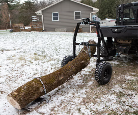 A Log Hauler, attached to an off-road utility vehicle, lifts one end of a large log off the snowy ground for transport. A house with trees and patchy snow is visible in the background.