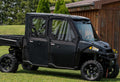 A Polaris Ranger 900/1000 Crew (2013-2018) with Solid Convertible Doors is parked on grass beside a wooden building, with evergreen trees, a lawn, and sunny skies in the background.