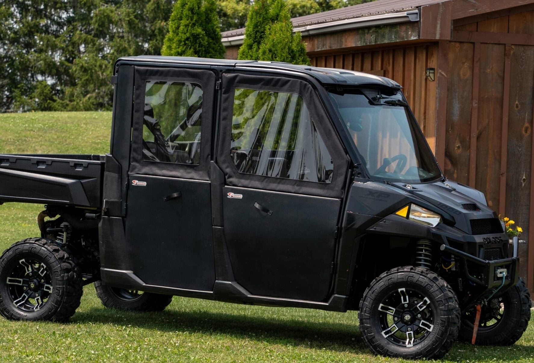 A Polaris Ranger 900/1000 Crew (2013-2018) with Solid Convertible Doors is parked on grass beside a wooden building, with evergreen trees, a lawn, and sunny skies in the background.
