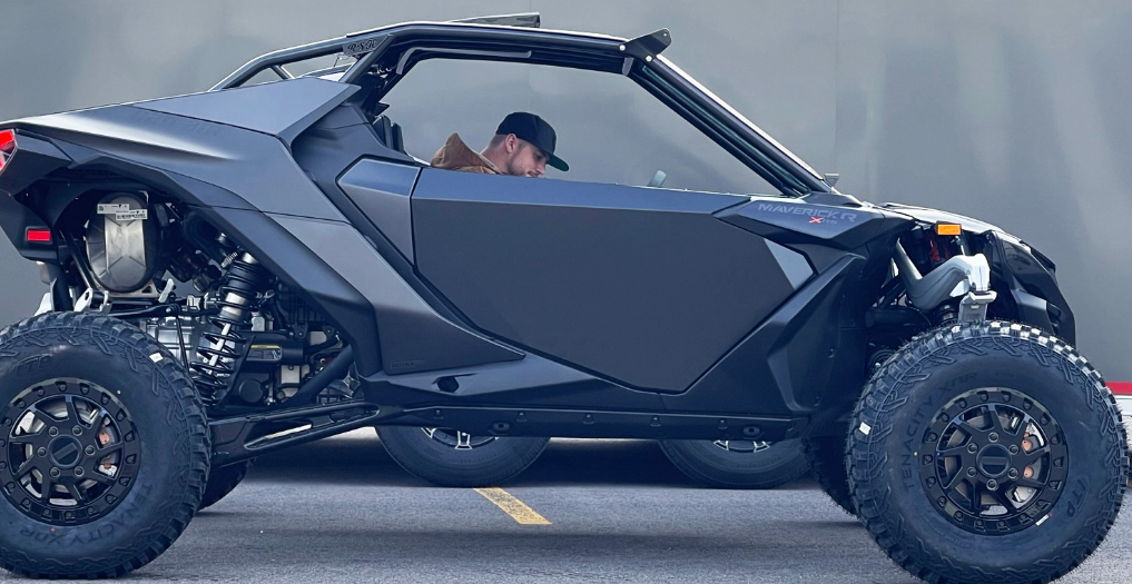 A man in a cap and brown jacket sits inside a matte black Can Am Maverick R fitted with the 2 Seat Door Set, featuring large rugged tires and parked on asphalt marked by a yellow line. The blurred background shows gray tones.