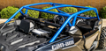 A close-up of a black Can-Am Maverick X3 with the S3 Power Sports 2017+ Custom Abc Pillar Cage in blue, bucket seats, minimalist dashboard, and steering wheel. Green bushes and a brown brick wall are visible in the background.