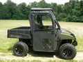 A Polaris Midsize Round Tube Ranger with large off-road tires is parked in a grassy field. It features the Framed Door Kit with clear plastic windows on the doors and roof. Green forest trees and brush can be seen behind it.