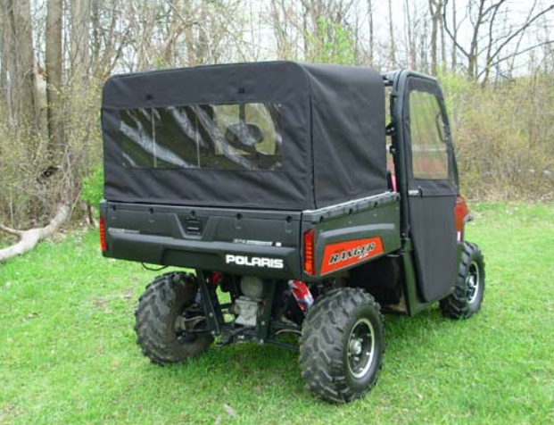 A black Polaris Ranger utility vehicle, equipped with the Polaris Ranger Midsize Soft Bed Cover Shell featuring clear side and rear windows, is parked on grass near leafless trees. The cover protects the cab and cargo area; the rear wheels are large and r