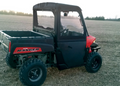 A Polaris Ranger with Midsize Convertible Doors, in red and black, is parked in a harvested field. It features a closed cab, rugged tires, and cargo bed, with trees and a pale sky in the background.