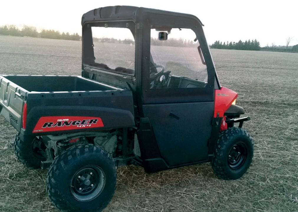 A Polaris Ranger Midsize with Pro-Fit Cage Convertible Doors and black and red accents is parked on a dry field at sunset, featuring a closed cab, rugged tires, and a cargo bed. Farmland and trees appear in the background beneath a pale sky.