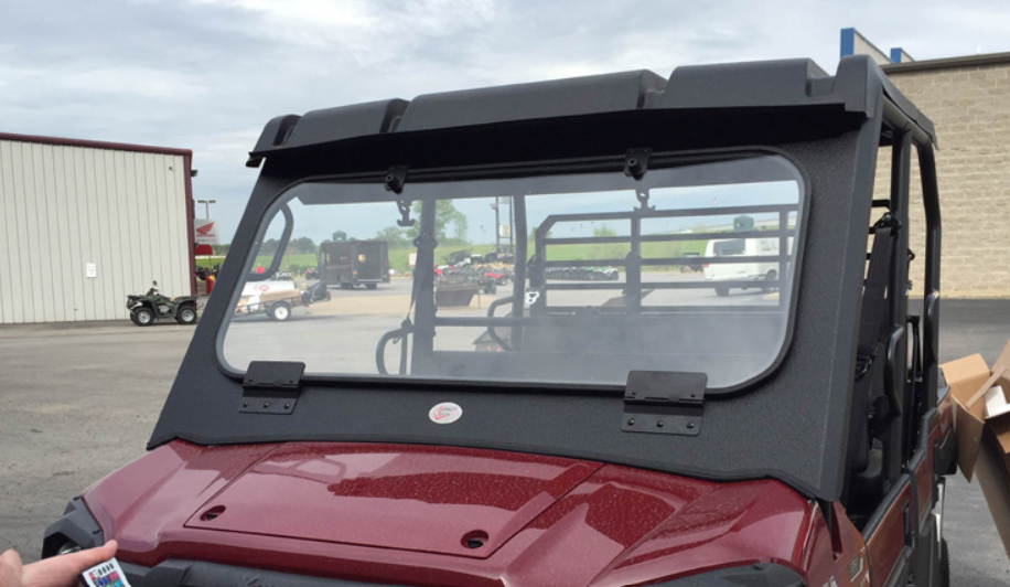 A Mule Pro FX/FXT/DX/DXT Fold Down Glass Windshield with a black frame and bottom hinges is mounted on a red utility vehicle parked outdoors. Clear glass, other vehicles, and buildings appear in the background under a cloudy sky.