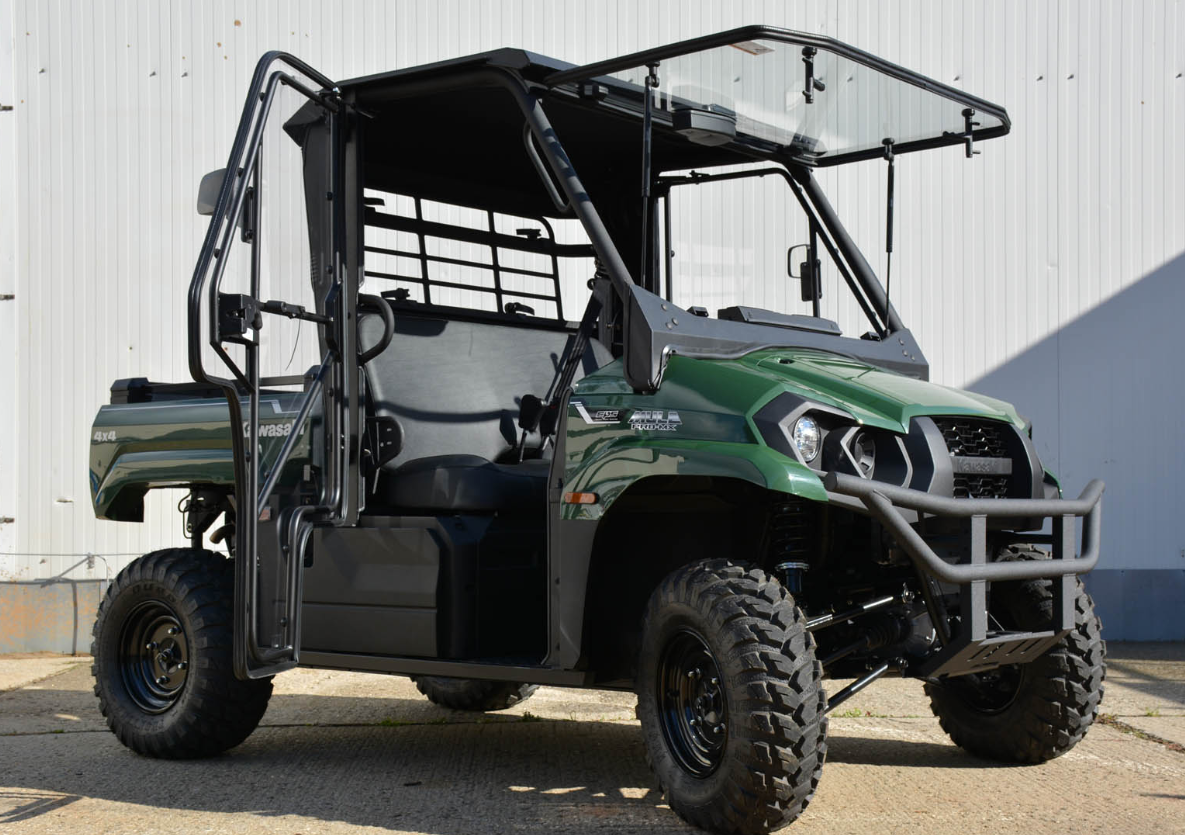 A Kawasaki Mule MX with the Cab Kit, four-wheel drive, open cab with side rails, black roof, protective front grill, rugged off-road tires, and seating for two is parked on concrete in front of a white metal building.