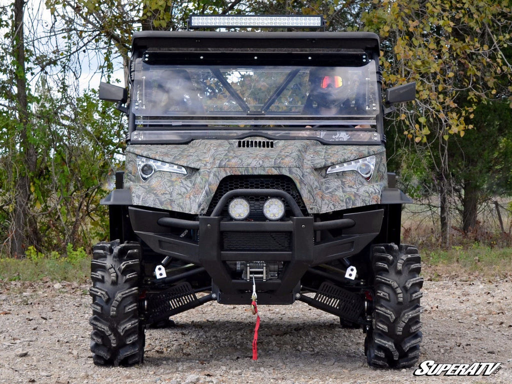 An ODES Dominator featuring a scratch-resistant flip windshield is parked on gravel, surrounded by trees. The rugged vehicle has camo patterns, deep-tread tires, LED headlights, a winch, and SuperATV branding visible in front.