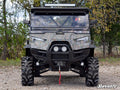 An ODES Dominator featuring a scratch-resistant flip windshield is parked on gravel, surrounded by trees. The rugged vehicle has camo patterns, deep-tread tires, LED headlights, a winch, and SuperATV branding visible in front.