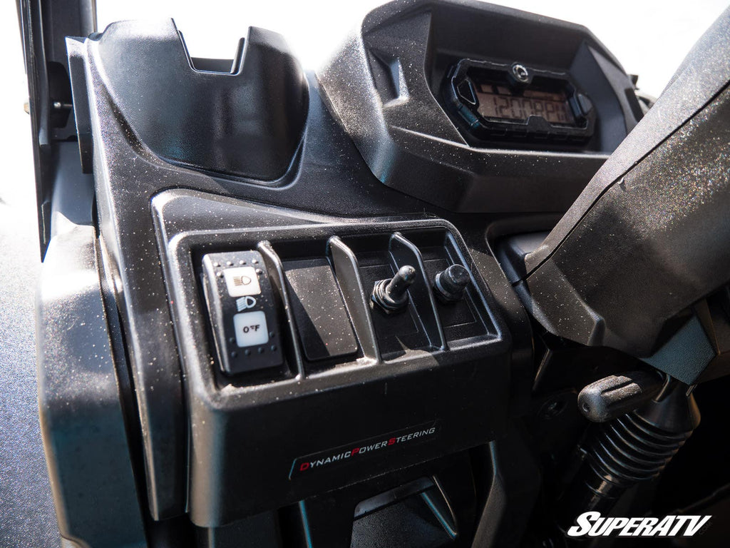 Close-up of a Can-Am Maverick X3 Plug & Play Turn Signal Kit installed on a vehicle dashboard with toggle switches, a button, and display screen labeled Dynamic Power Steering. The dusty surface features the SuperATV logo in the bottom right corner.