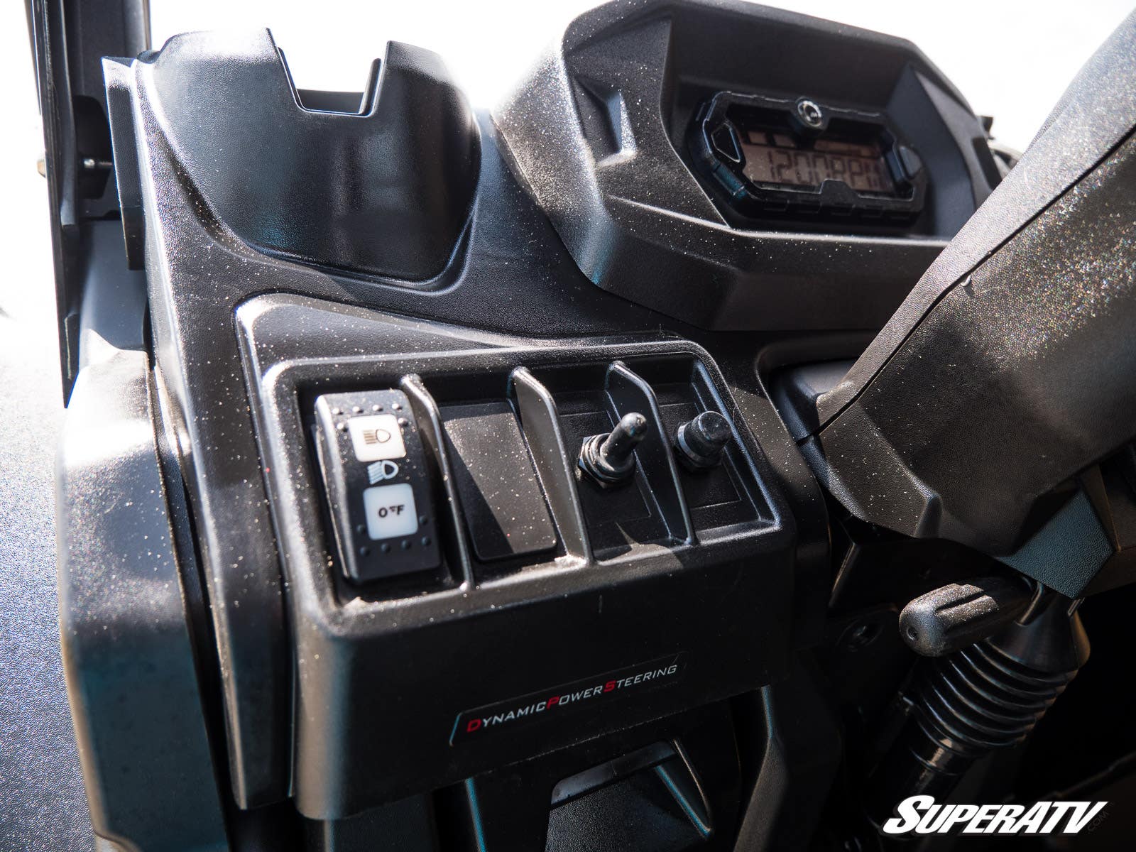 Close-up of a Can-Am Maverick X3 Plug & Play Turn Signal Kit installed on a vehicle dashboard with toggle switches, a button, and display screen labeled Dynamic Power Steering. The dusty surface features the SuperATV logo in the bottom right corner.