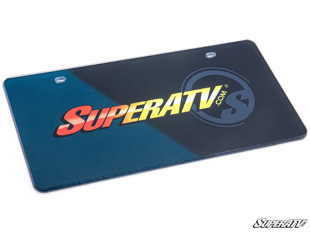 The SuperATV Front Vanity Plate features a dark blue and black diagonal background, bold “SUPERATV.COM” text, a faint circular logo, two top mounting holes, and the SuperATV logo in the bottom right corner.