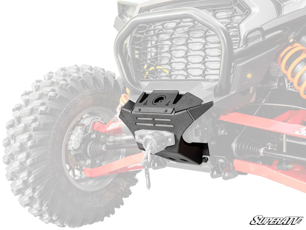 A close-up of a Polaris RZR XP Winch Mount, black and heavy-duty, installed on the front of an off-road vehicle. The centrally positioned mount shows bolts and a cable hole, with faded tires and suspension. “SuperATV” logo appears in the bottom right corn