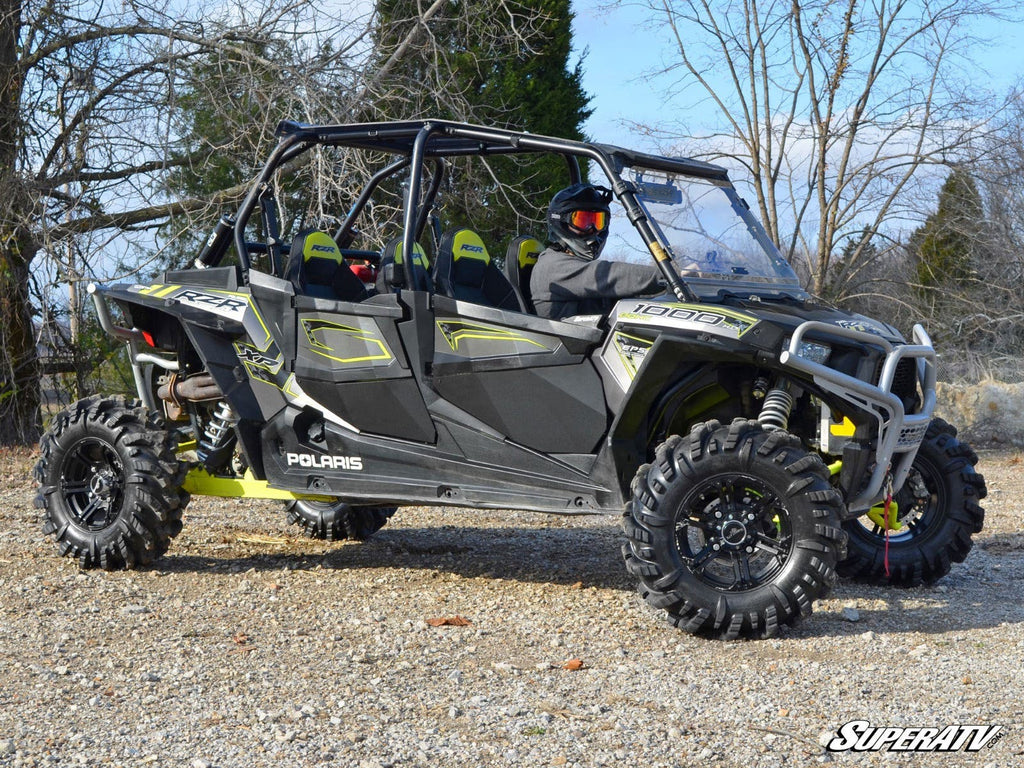 A black and green Polaris RZR 4 900 UTV with SuperATV lower doors and large off-road tires is parked on gravel. Four seats are visible; a helmeted driver sits up front. Bare trees and a cloudy sky fill the background; “SuperATV” appears in the corner.