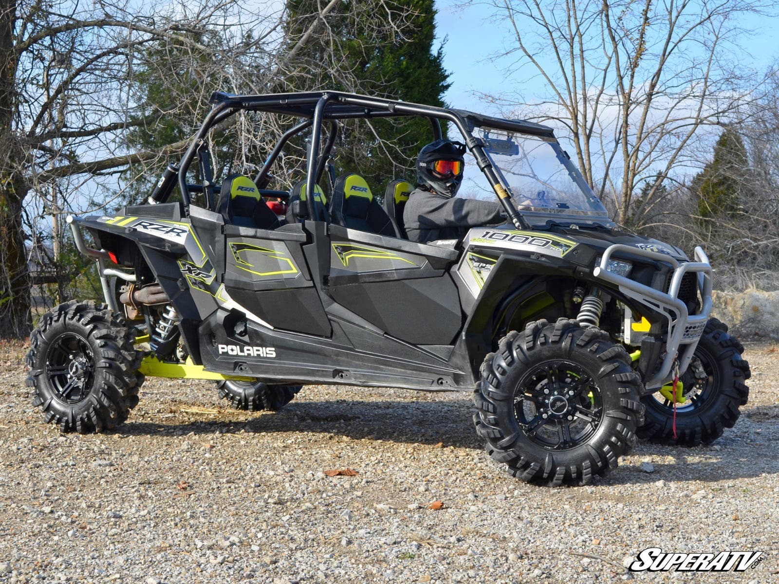 A black and green Polaris RZR 4 900 UTV with SuperATV lower doors and large off-road tires is parked on gravel. Four seats are visible; a helmeted driver sits up front. Bare trees and a cloudy sky fill the background; “SuperATV” appears in the corner.