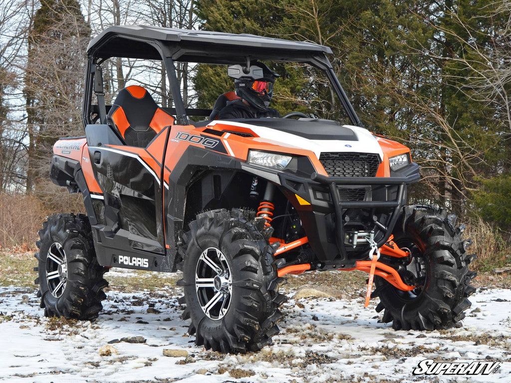 A Polaris side-by-side equipped with a Polaris General 3 Lift Kit and large tires is parked on patchy snow. The helmeted driver wears red goggles, leafless trees stand in the background, and the SuperATV logo appears in the corner.