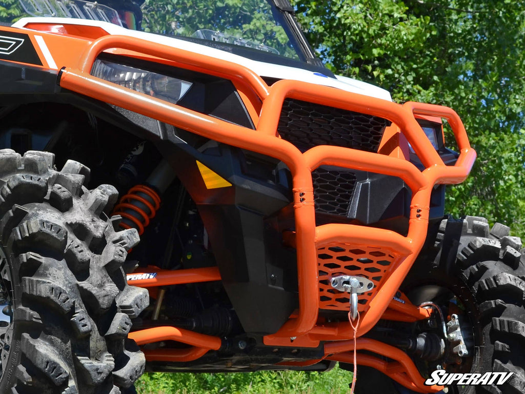 Close-up of a Polaris General Front Bumper with large knobby tires, an orange grille guard, and suspension springs. Parked outdoors amid green trees, the SuperATV logo appears in the bottom right corner.