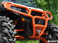 Close-up of a Polaris General Front Bumper with large knobby tires, an orange grille guard, and suspension springs. Parked outdoors amid green trees, the SuperATV logo appears in the bottom right corner.