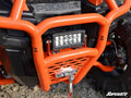 Close-up of a Polaris General XP 1000 Front Bumper, showing an orange finish, rectangular LED light bar, metal winch with hook, mesh grille, rugged tire, thick protective bars, and the SuperATV logo in the bottom right.