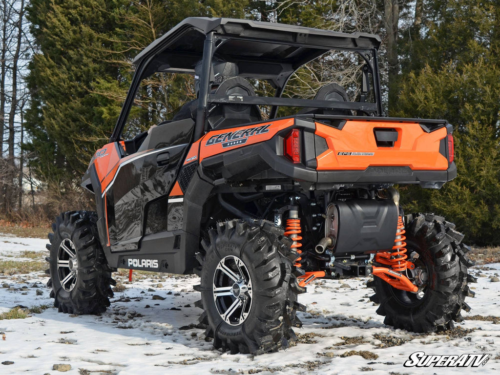 A Polaris General 1000 UTV equipped with Polaris General High Clearance 1.5 Rear Offset A-Arms is parked on snowy ground, featuring large off-road tires and orange suspension springs, near evergreen and leafless trees.