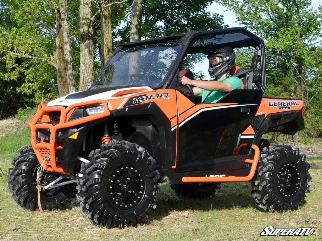A rider in a helmet and green shirt drives an orange and black Polaris General with large tires on grassy terrain. Displayed is the Polaris General Front Leveling Kit. The SuperATV logo appears in the lower right corner against a backdrop of trees.