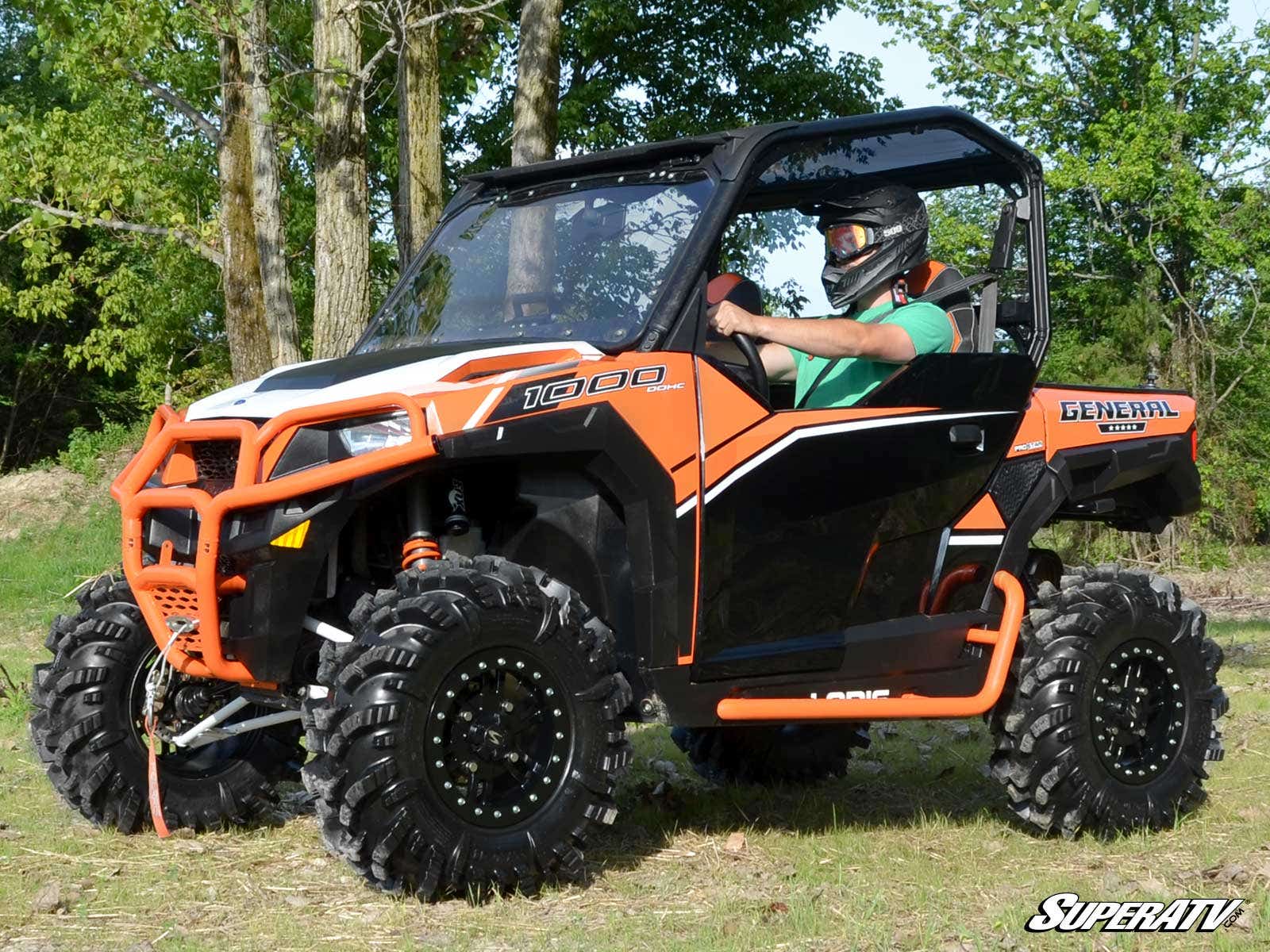 A rider in a helmet and green shirt drives an orange and black Polaris General with large tires on grassy terrain. Displayed is the Polaris General Front Leveling Kit. The SuperATV logo appears in the lower right corner against a backdrop of trees.