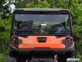 Rear view of a Polaris General XP 1000 with the SuperATV Rear Windshield, black roof, and two headrests. The vehicle is set among lush green trees. The SuperATV logo is visible in the bottom right corner.